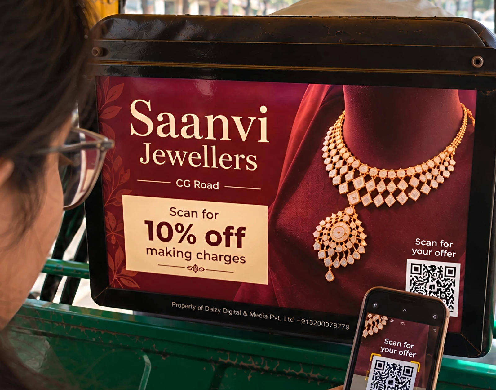 A lit board in an auto showing a sample jewellery shop ad with a 10% off offer and a QR code, which a passenger is scanning with her phone.